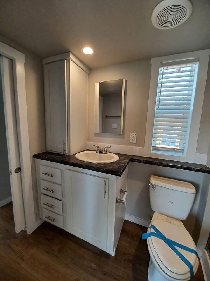 Modern bathroom with white cabinetry, a dark countertop, and a round sink. A mirror is above. The toilet has blue tape and a window blind filters light.
