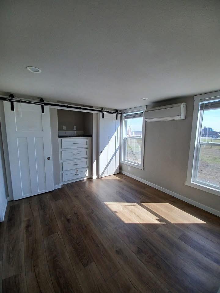 Bright room with wooden floors, large windows, and an air conditioner. White sliding barn doors reveal built-in drawers, creating a minimalist feel.