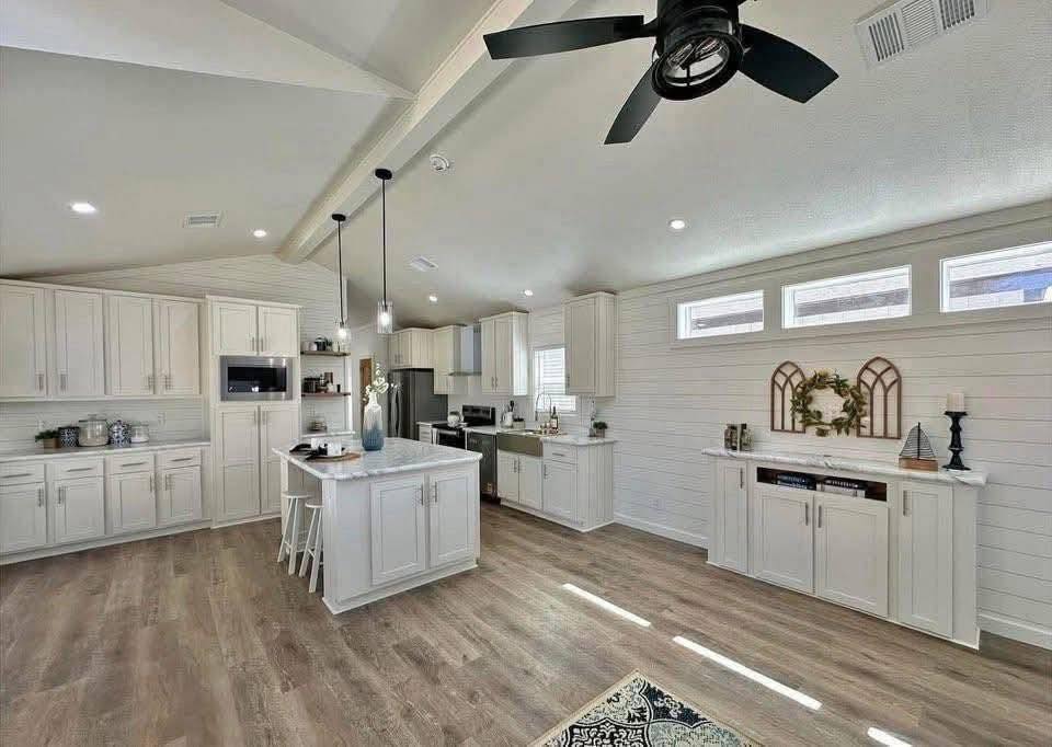 Spacious modern kitchen with white cabinets, central island, and wood flooring. Natural light streams through upper windows. Ceiling fan and pendant lights add elegance.