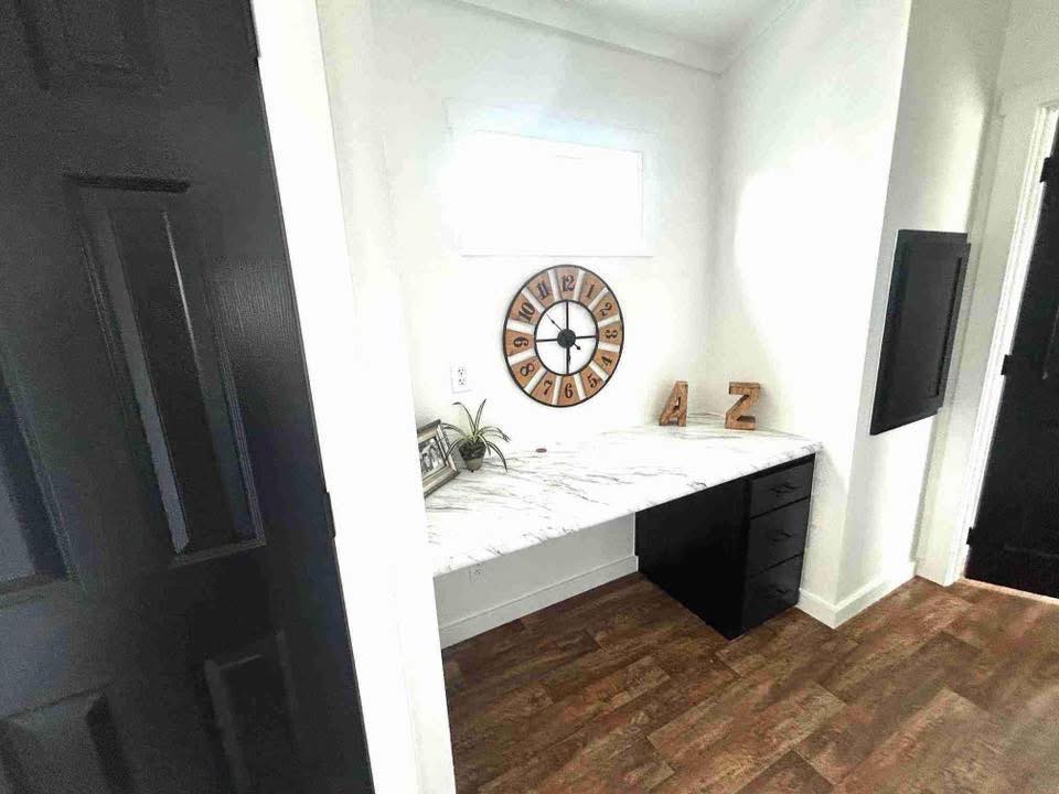 Minimalist home office nook with a marble desk, black drawers, large wall clock, small plant, and decorative letters. Wood flooring and white walls.