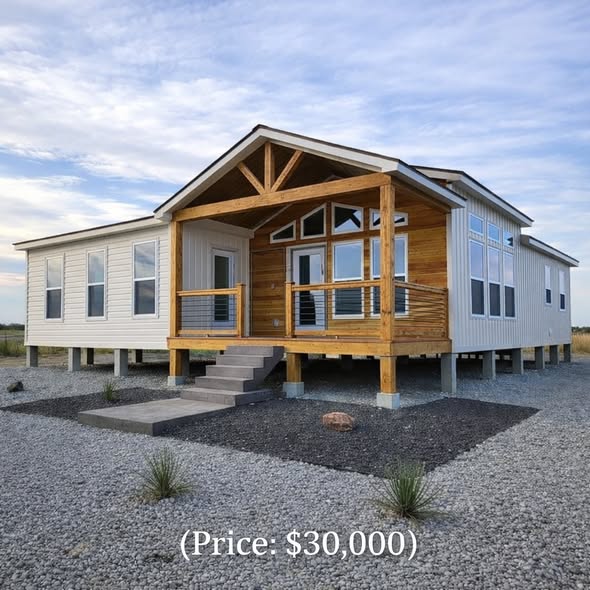 Modern manufactured home on a gravel lot, featuring large windows and a wooden front porch under a clear sky. Price is shown as $30,000.