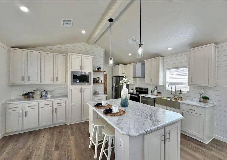 Spacious kitchen with light wooden cabinets, an island with stools, marble countertops, pendant lights, and decorative plants, conveying elegance.
