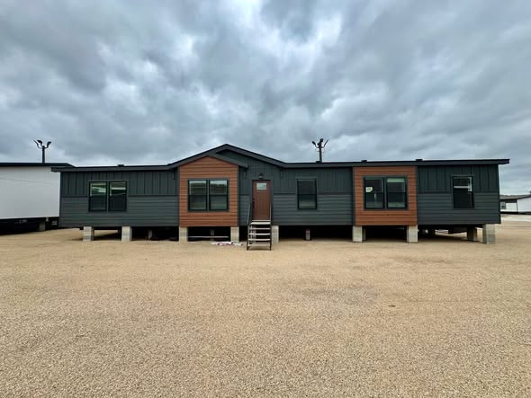 Modern, dark-colored modular home on a gravel lot under a cloudy sky. The structure is elevated on concrete supports, conveying a minimalist design.