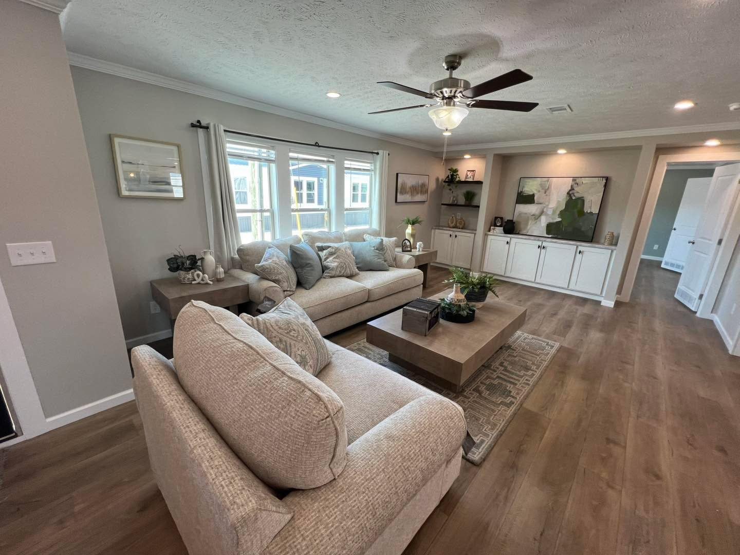 Spacious living room with beige sofas, wooden coffee table, ceiling fan, and decor on built-in shelves. Sunlight filters through large windows. Cozy and modern.