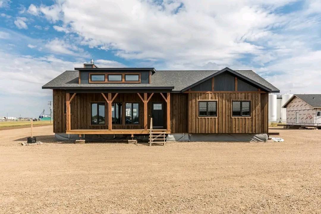 A rustic, wooden cabin with large windows and a gabled roof sits on a gravel lot under a partly cloudy sky, conveying a sense of openness and simplicity.