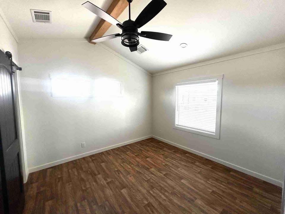 A bright, empty room with white walls, a wood beam ceiling, and wood flooring. Features a black ceiling fan, two small windows, and a larger window with blinds.