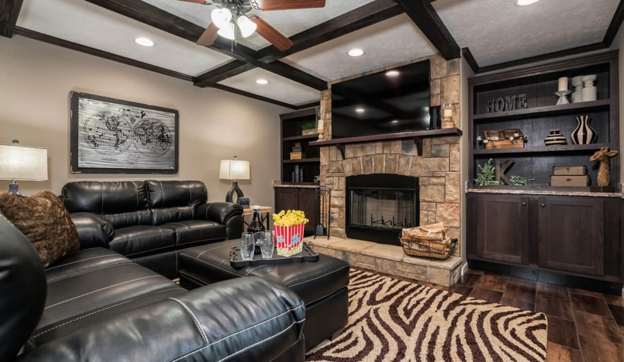 Cozy living room with dark leather sofas, stone fireplace, and large TV. Zebra-pattern rug and wood beams add warmth. Bookshelves hold decor items.
