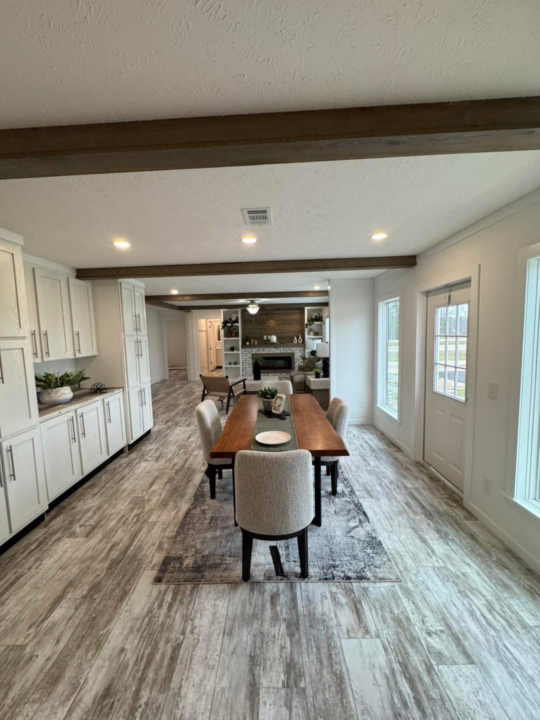 Open concept dining area with a wooden table, six upholstered chairs, and large windows. White cabinetry lines the left wall, leading to a cozy living space.