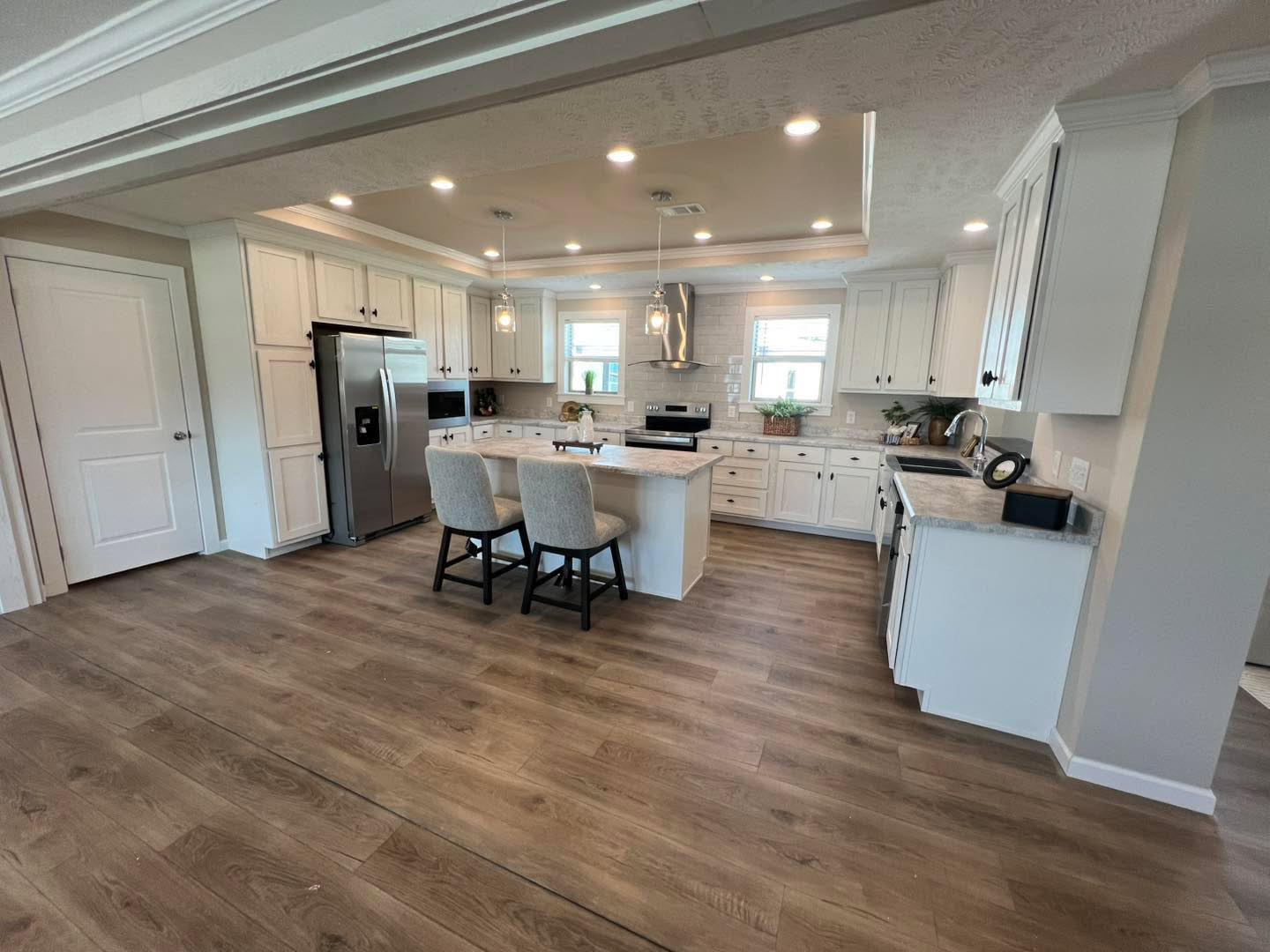 Spacious kitchen with white cabinets, stainless steel appliances, and a central island with two chairs. Warm wood floors and bright lighting create an inviting atmosphere.