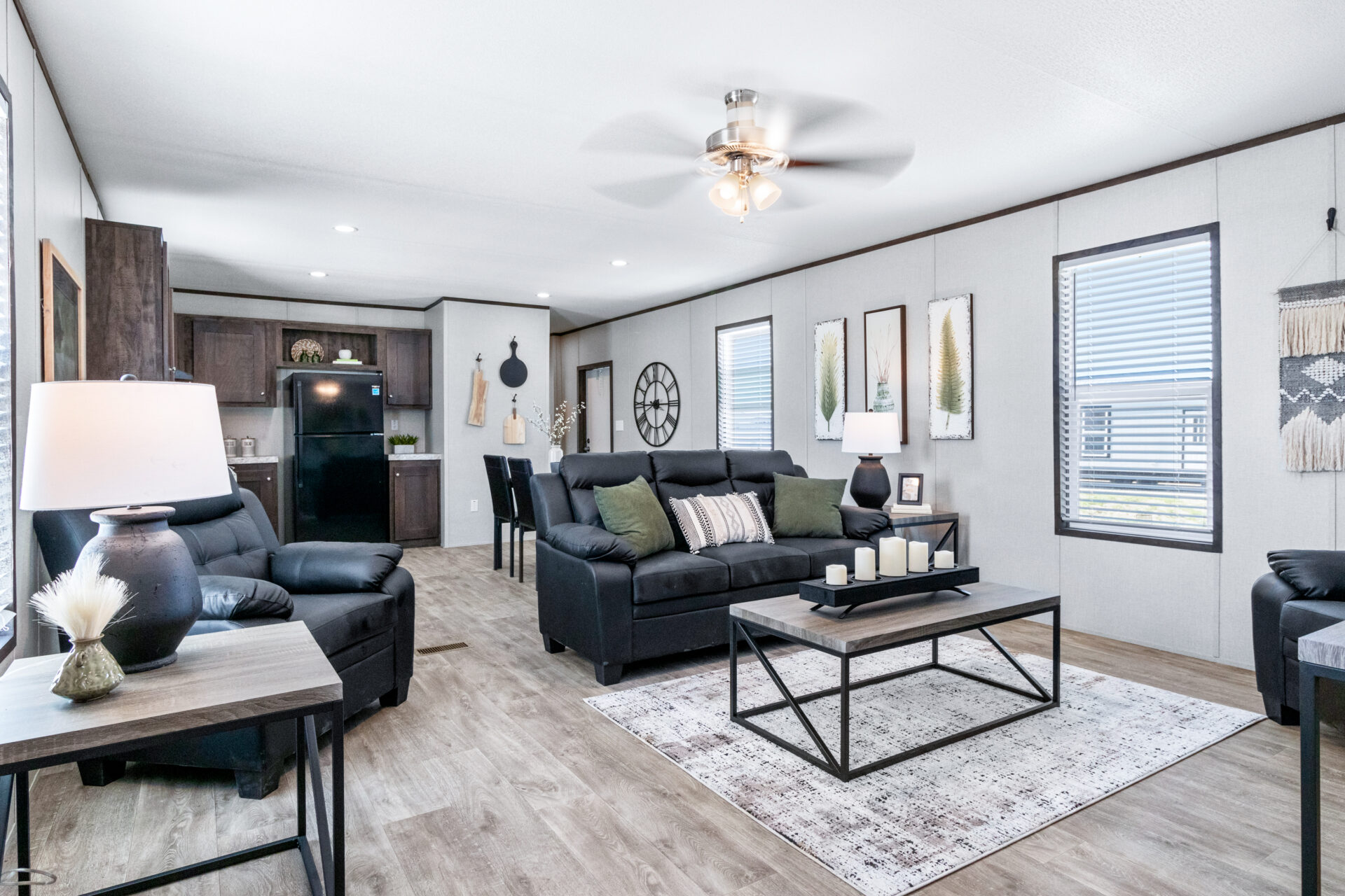 Modern living room with dark sofas, green and striped cushions, and a black coffee table with candles. Wooden floor, textured wall decor, and ceiling fan enhance coziness.