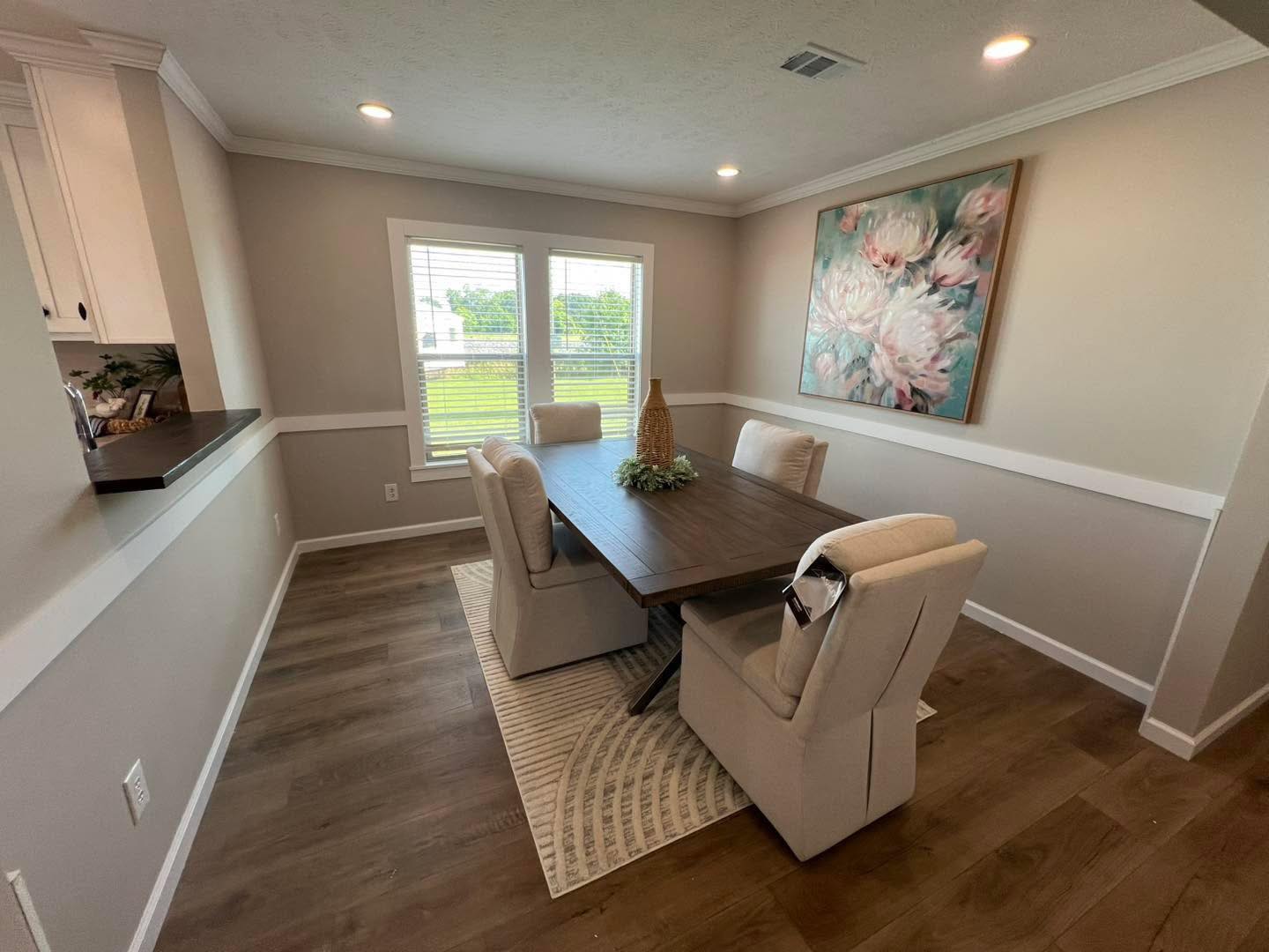 Elegant dining area with a dark wooden table, beige cushioned chairs, and floral artwork. Sunlight streams through a window, illuminating the cozy space.