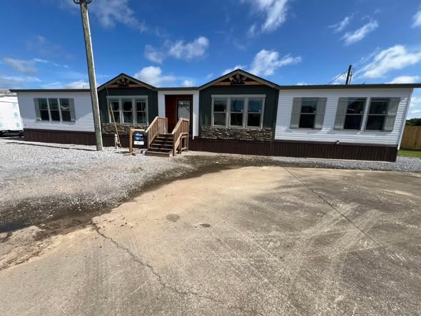 Single-story manufactured home with white siding and stone accents, featuring large windows and a wooden porch. Sunny day with blue sky and scattered clouds.