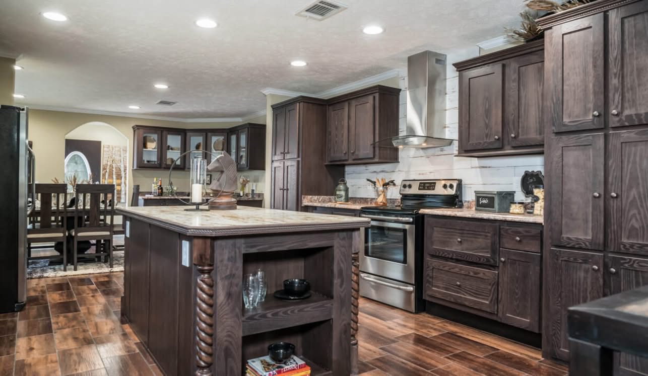 A warm, rustic kitchen with dark wooden cabinets and stainless steel appliances. A central island features intricate woodwork. Dining area visible with soft lighting.