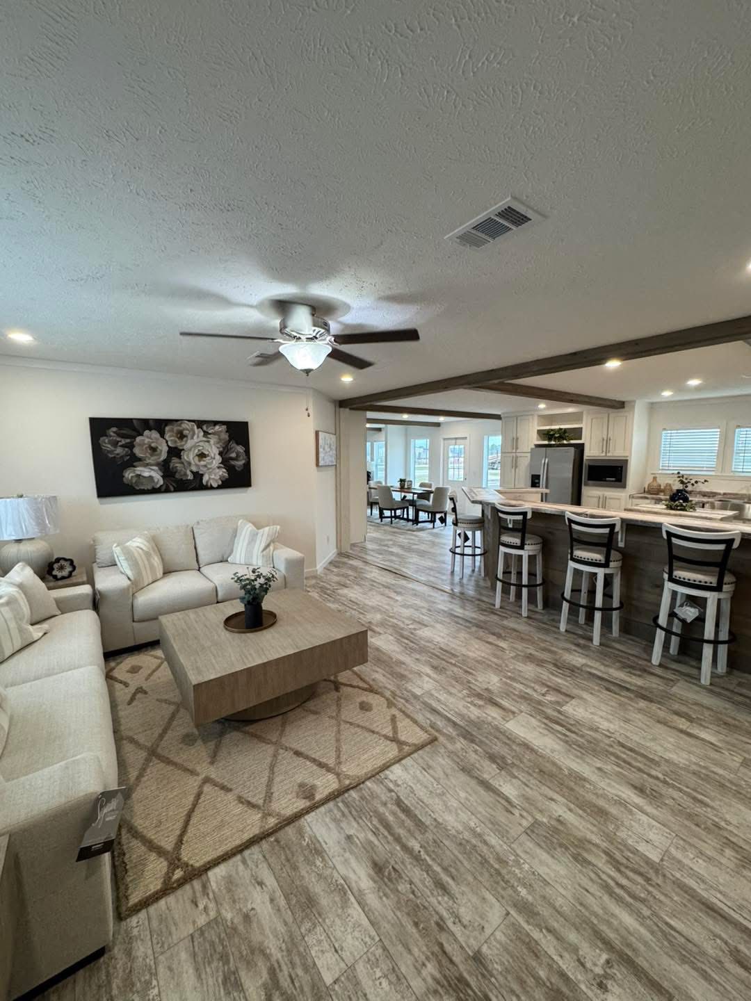 Spacious open-plan living area with light wood floors, a cozy sectional sofa, floral artwork, and a large kitchen island with stools. Bright and inviting atmosphere.