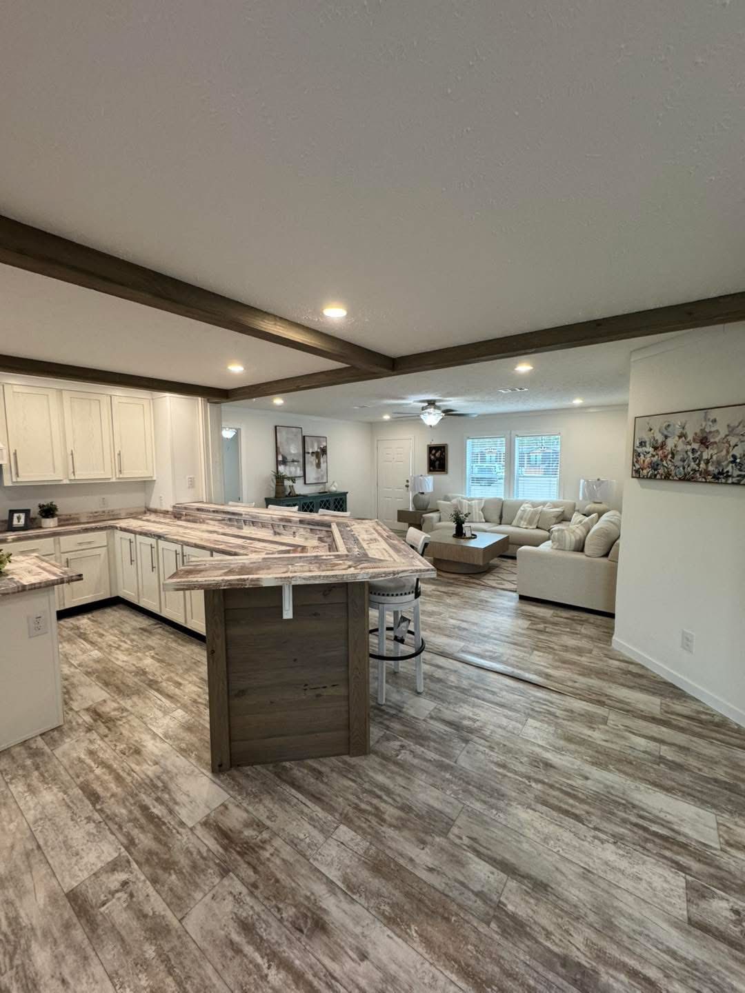 Spacious open-plan kitchen and living area with wooden floors. Features a large kitchen island, white cabinets, a cozy beige sofa, and bright natural light.