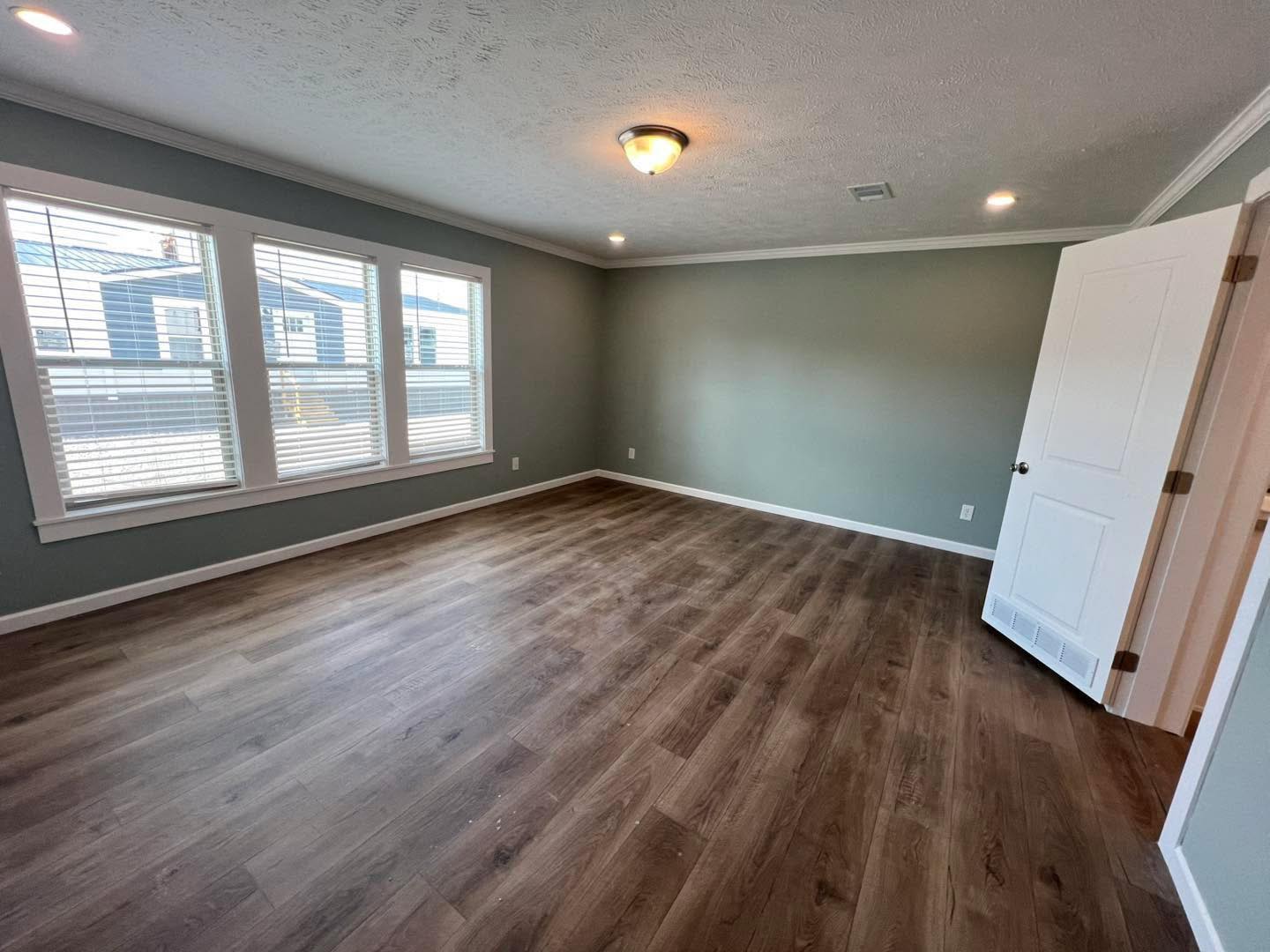 Spacious room with gray walls, wooden floor, and a ceiling light. Three large windows allow natural light, and an open white door adds a welcoming feel.