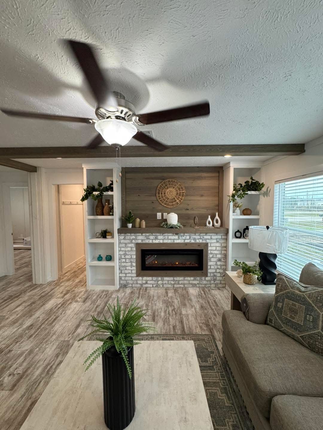 Cozy living room with a ceiling fan and electric fireplace on a brick wall. Open shelves display plants and decor, creating a warm, inviting ambiance.