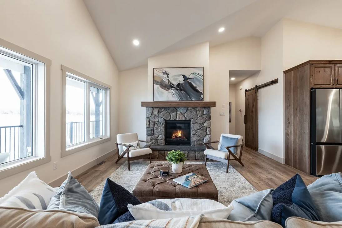 Cozy living room with stone fireplace, abstract art, soft chairs, and a plush ottoman. Sunlight streams through large windows, creating a warm, inviting ambiance.