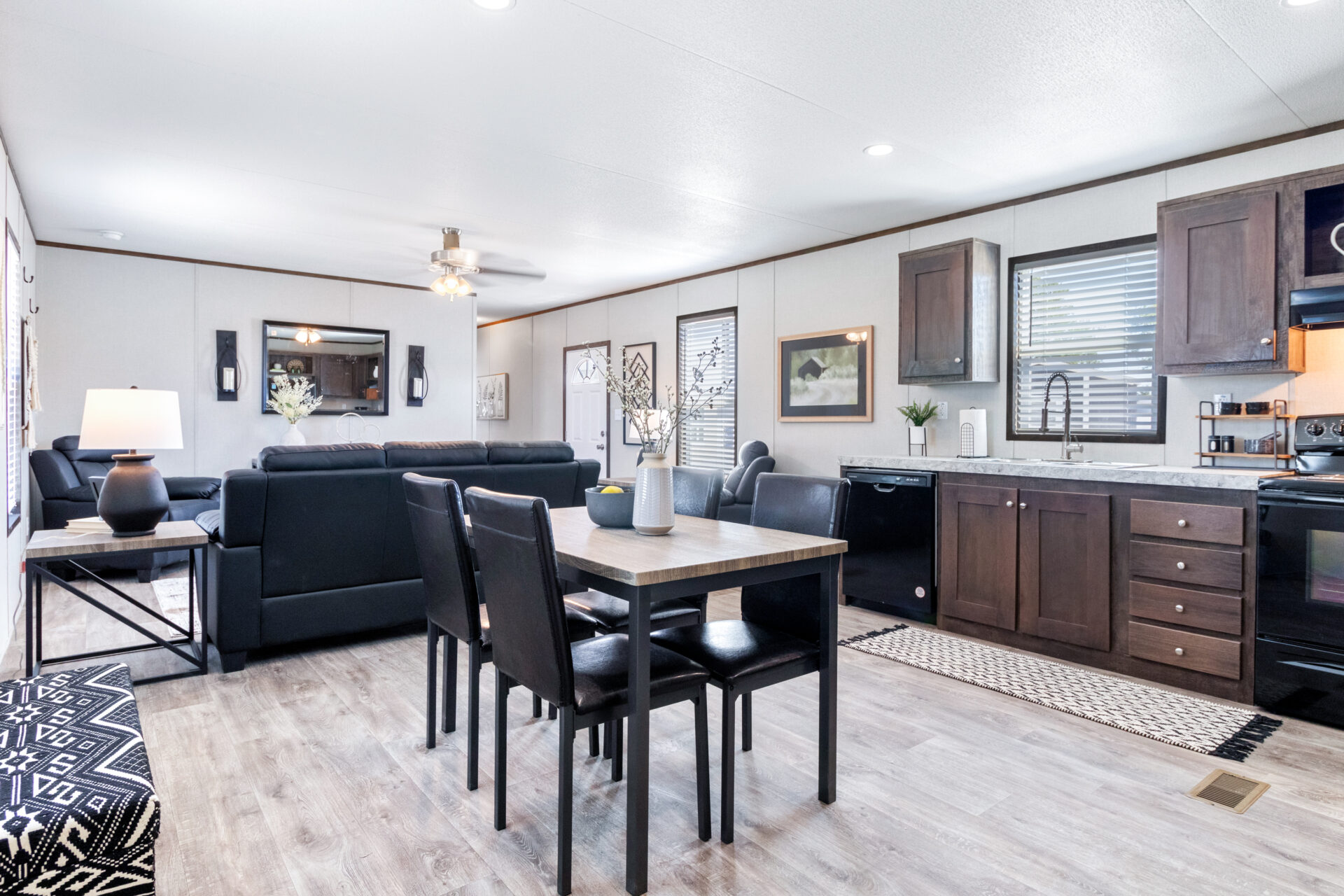 Open-concept living space with a dining table and four chairs in the center. Nearby, a modern kitchen features dark wood cabinets. A cozy seating area with black sofas is in the background. Natural light fills the room, creating a welcoming atmosphere.