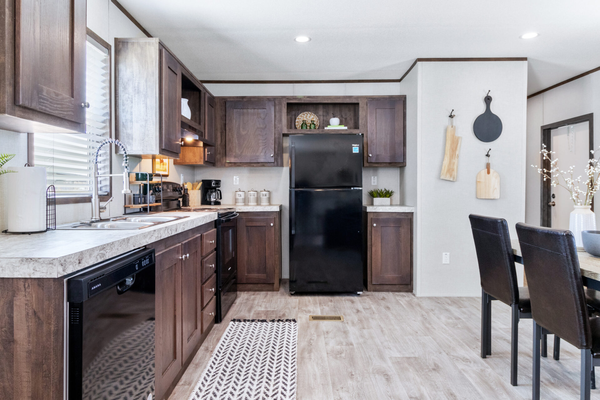 Modern kitchen with dark wood cabinets, black fridge, and dishwasher. Light wood floors, cutting boards on wall, dining table with black chairs. Cozy ambiance.