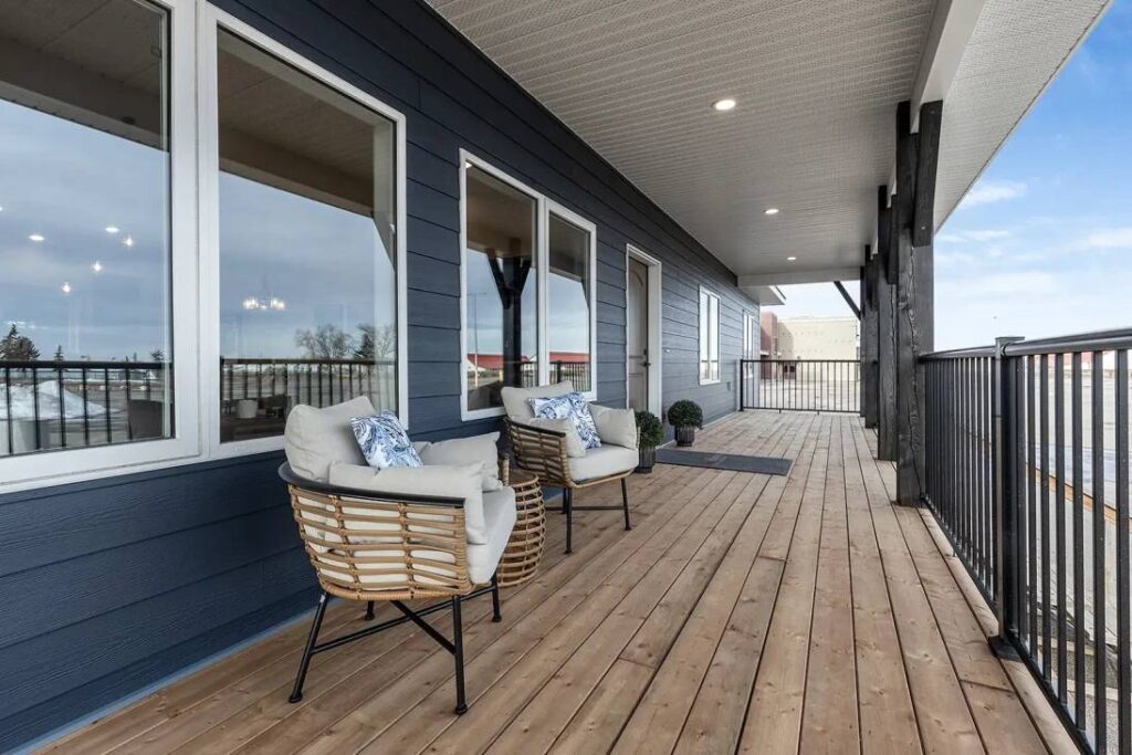 Spacious wooden balcony with two cushioned wicker chairs and a potted plant against a modern blue-paneled house. Bright, calming atmosphere.