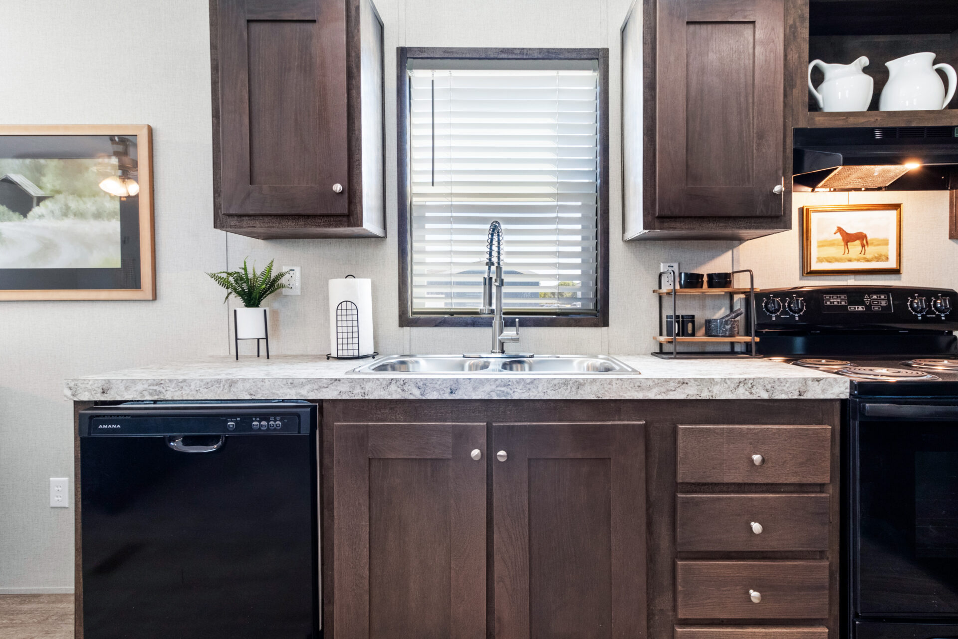 Modern kitchen with dark wood cabinets, marble countertop, and stainless steel sink. A window with blinds is above the sink, and decor includes a plant, framed art, and white pitchers.