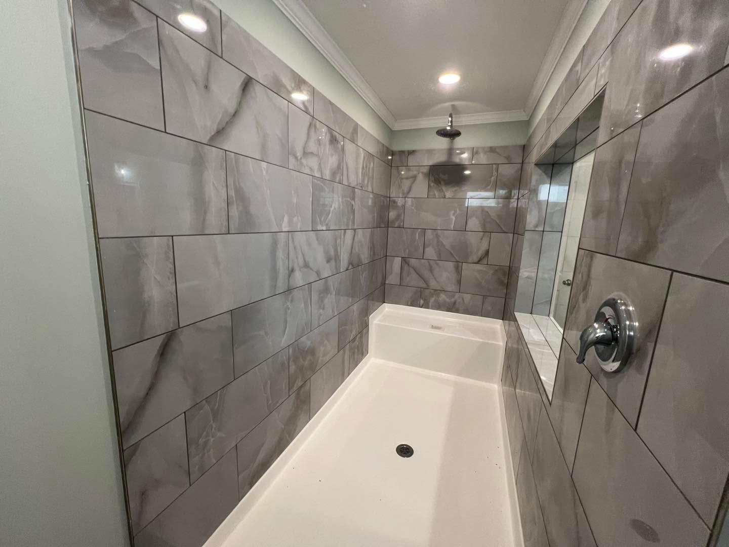 Modern shower with gray marble tiles, a large overhead showerhead, recessed lighting, and a seamless glass wall. The ambiance is sleek and calming.