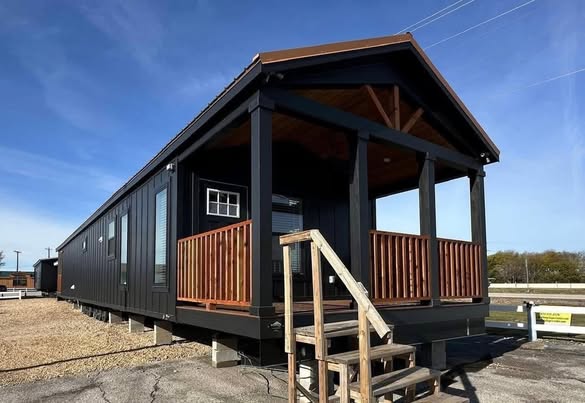 A modern, black tiny house with a wooden porch and stairs sits on a gravel lot under a clear blue sky, conveying a minimalist and cozy feel.