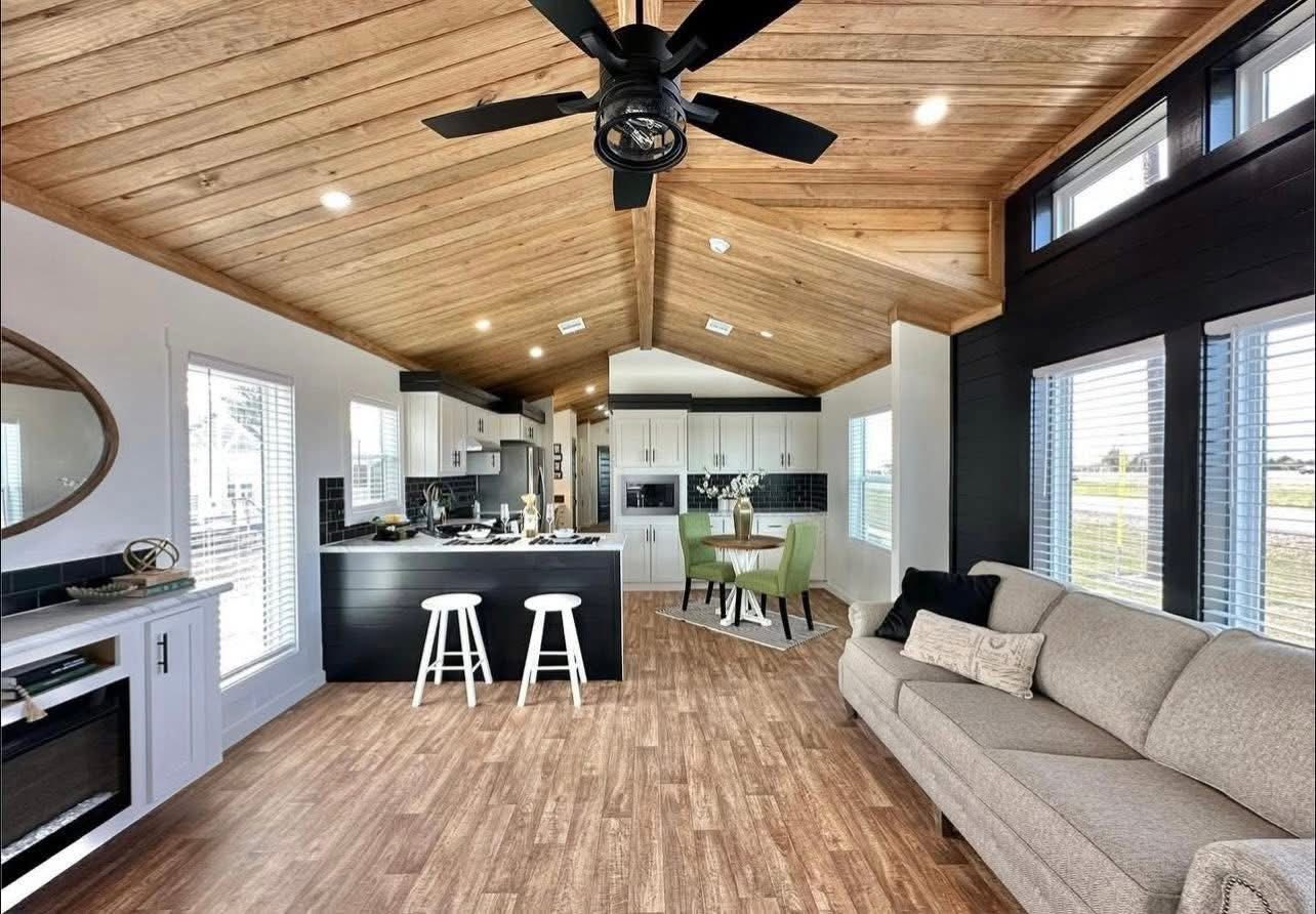 Cozy open-plan living area with wooden vaulted ceiling, modern kitchen, gray sofa, island with stools, and dining area with green chairs. Warm, inviting ambiance.