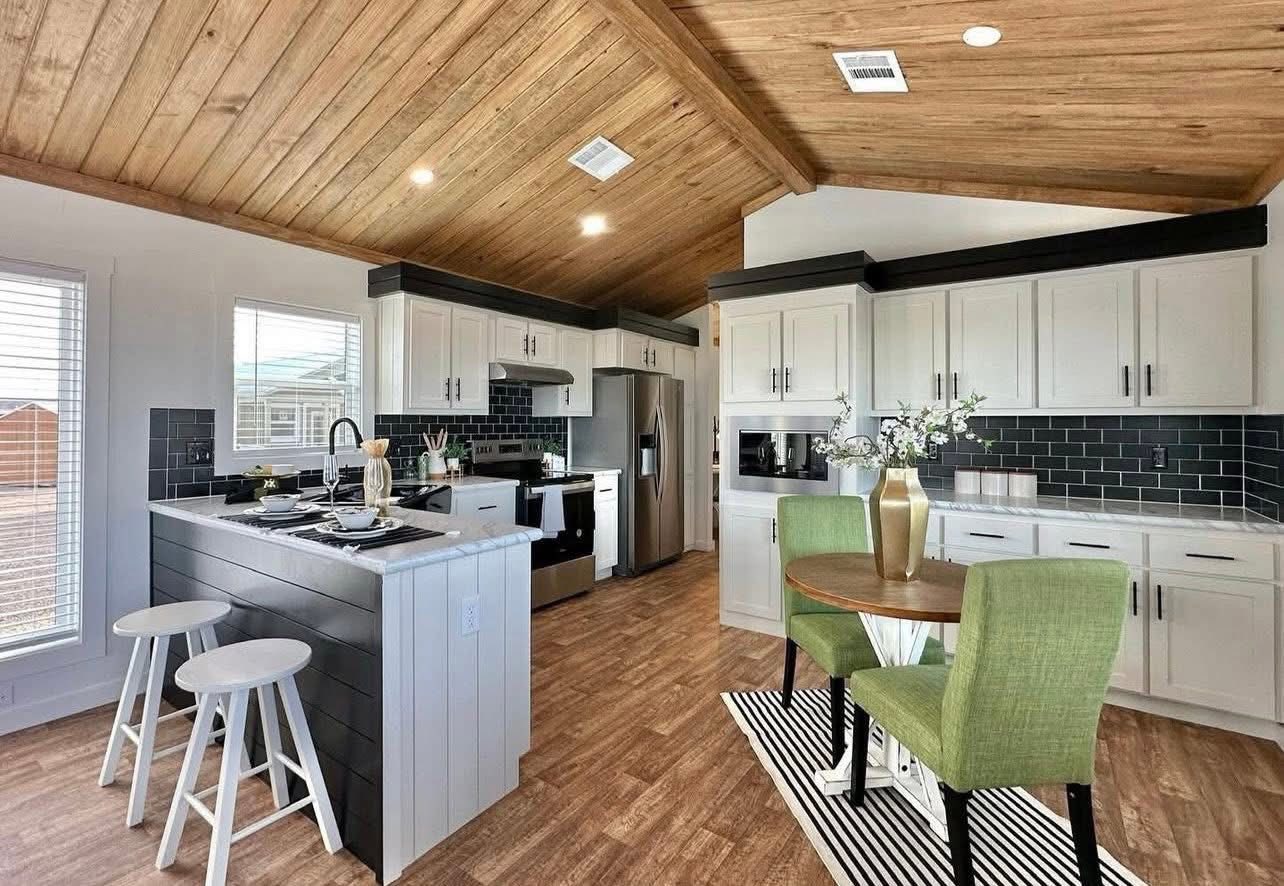 Modern kitchen with a wooden ceiling and floor, featuring white cabinets, a stainless steel fridge, and a black tile backsplash. A round table with green chairs and a centerpiece adds a cozy touch.
