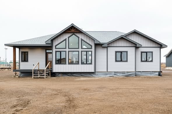 A modern, single-story house with a light gray exterior, triangular roof, large front windows, and a small porch, set in a barren landscape.