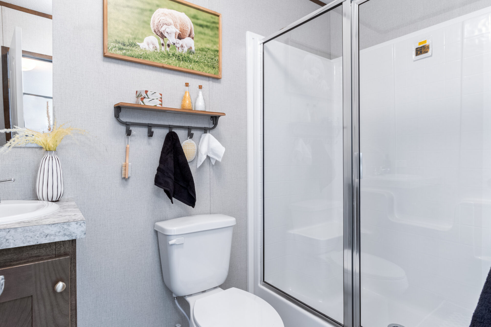Compact modern bathroom with a glass shower, toilet, and gray vanity. A shelf holds decor, and a sheep picture hangs above, adding a cozy touch.