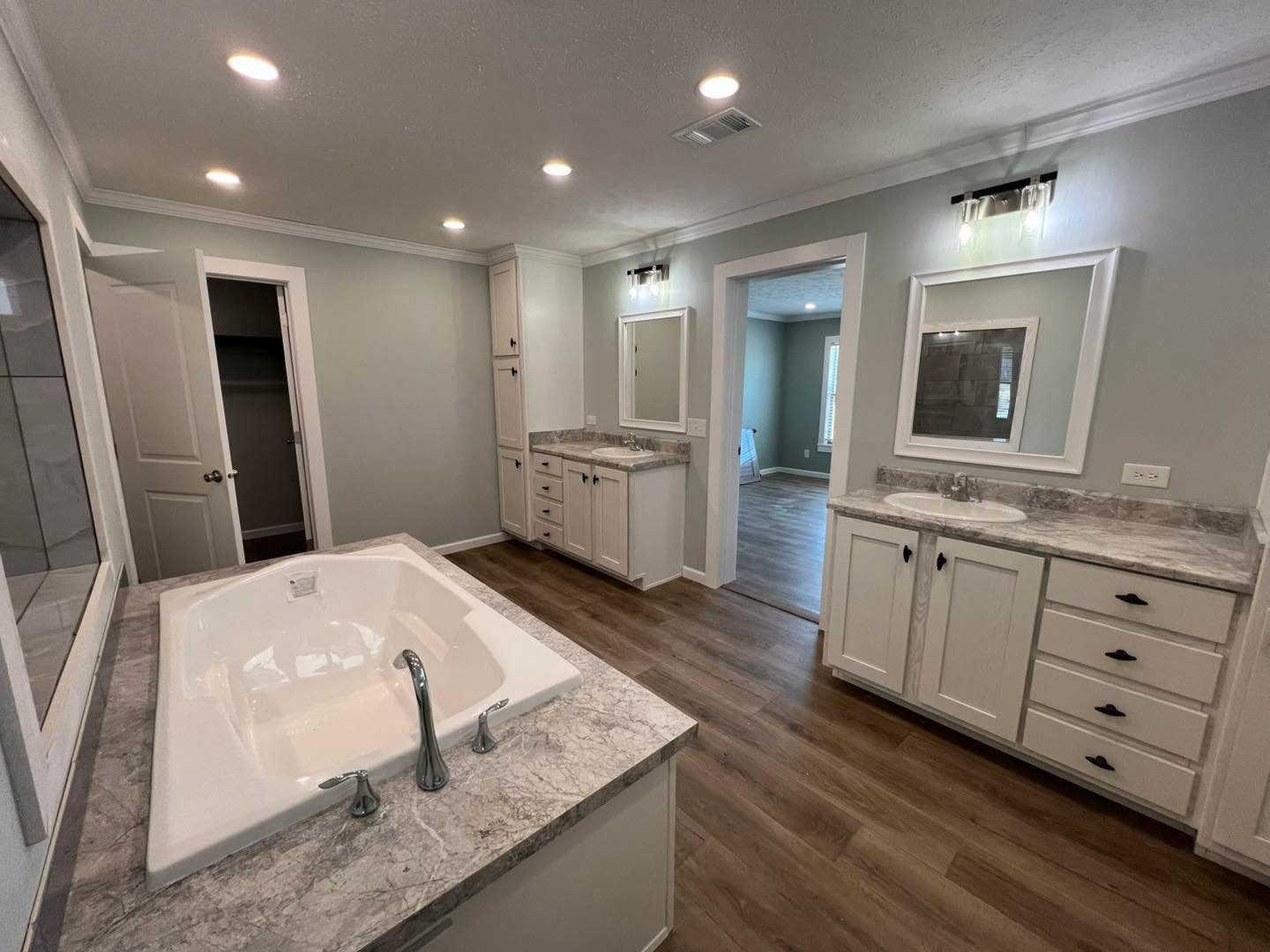 Spacious bathroom with a central marble bathtub, dual sinks with mirrors, and wood flooring. Bright, clean, and modern design evokes tranquility.