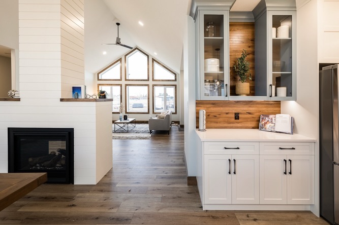 Modern open living space with high ceiling, large triangular windows, and wooden floor. White cabinetry and fireplace add a cozy, airy feel.