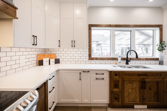 A modern kitchen with white cabinets, subway tile backsplash, and a wooden lower cabinet. A large window above the sink adds brightness.