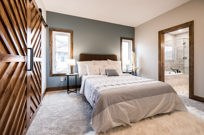 A cozy bedroom with a double bed, white and gray bedding, and a wooden headboard. There are two windows, a wooden sliding door, and an ensuite bathroom visible.