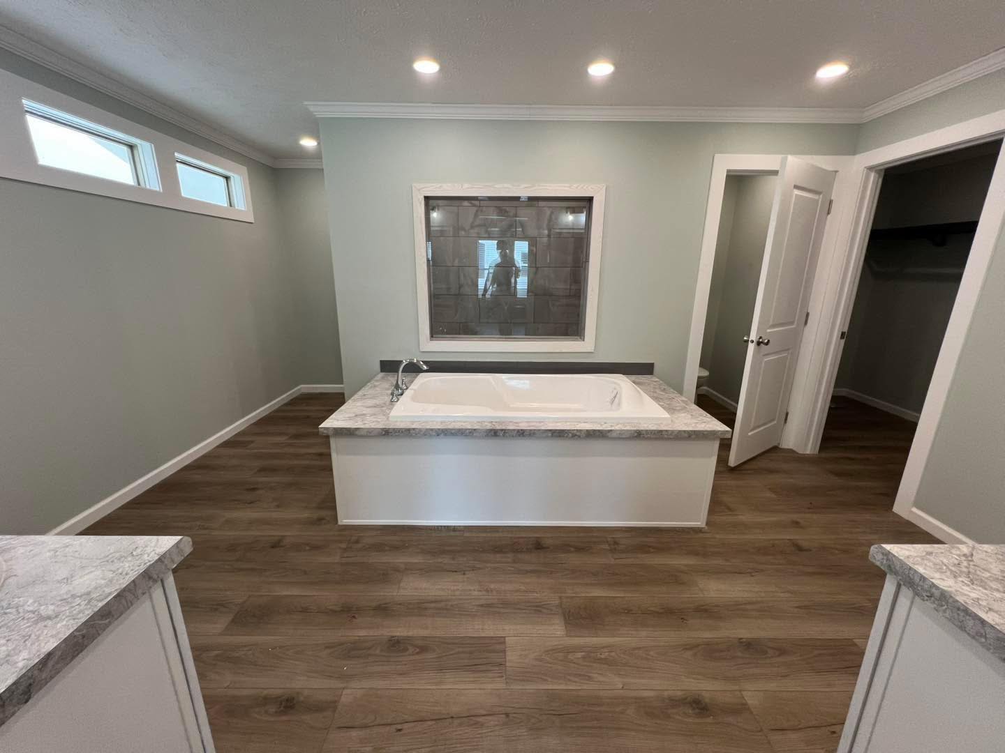 Spacious bathroom with a central white tub set in marble, wooden flooring, soft gray walls, recessed lighting, and a window with frosted glass.