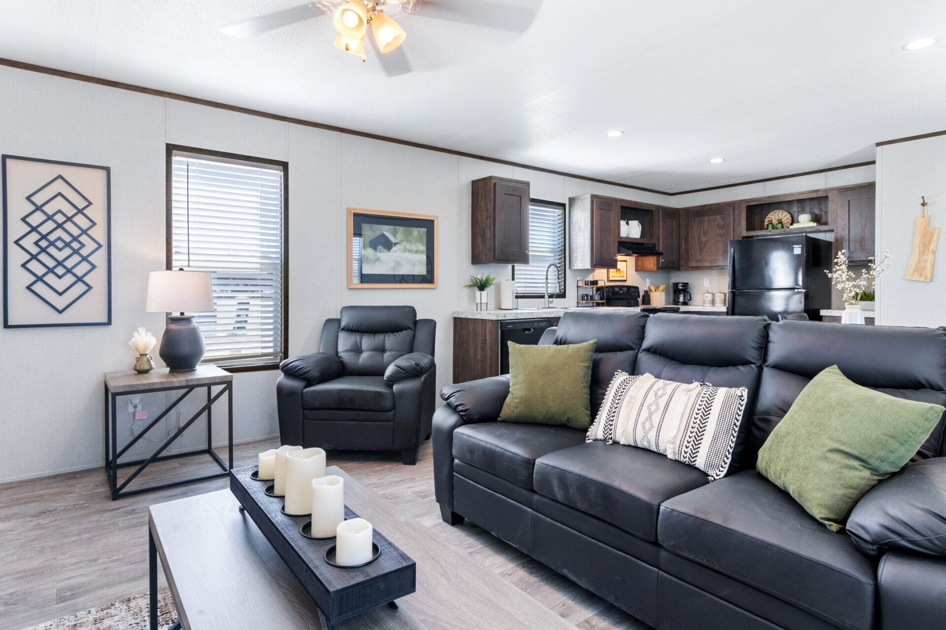 Modern living room with black leather sofas, green and patterned pillows, a coffee table with candles, and a view into a cozy kitchen with dark cabinets.