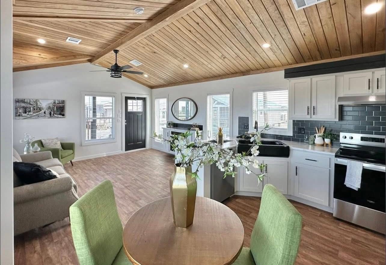 A cozy, modern open-plan living area with a wooden ceiling and floors. Features a kitchen with white cabinets, black backsplash, and a round dining table with green chairs, creating a warm, inviting atmosphere.