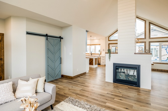 A spacious living room with light wood flooring features a cozy gray couch and pillows, a blue sliding barn door, and a modern fireplace under high ceilings.