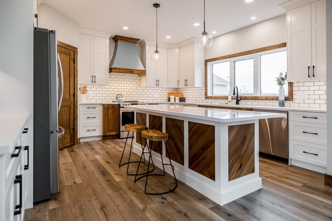 Modern kitchen with white cabinets, wooden accents, and a large island featuring two stools. Pendant lights hang from the ceiling, and a window provides natural light.