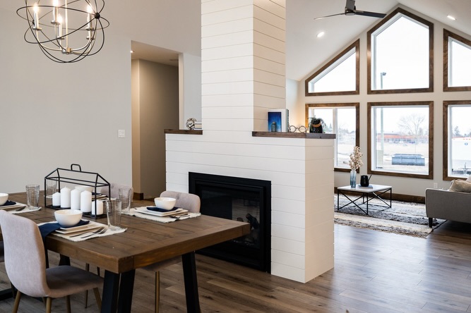 Modern open-concept living and dining area with a wooden table, candles, shiplap fireplace, large windows, a gray couch, and a stylish light fixture.