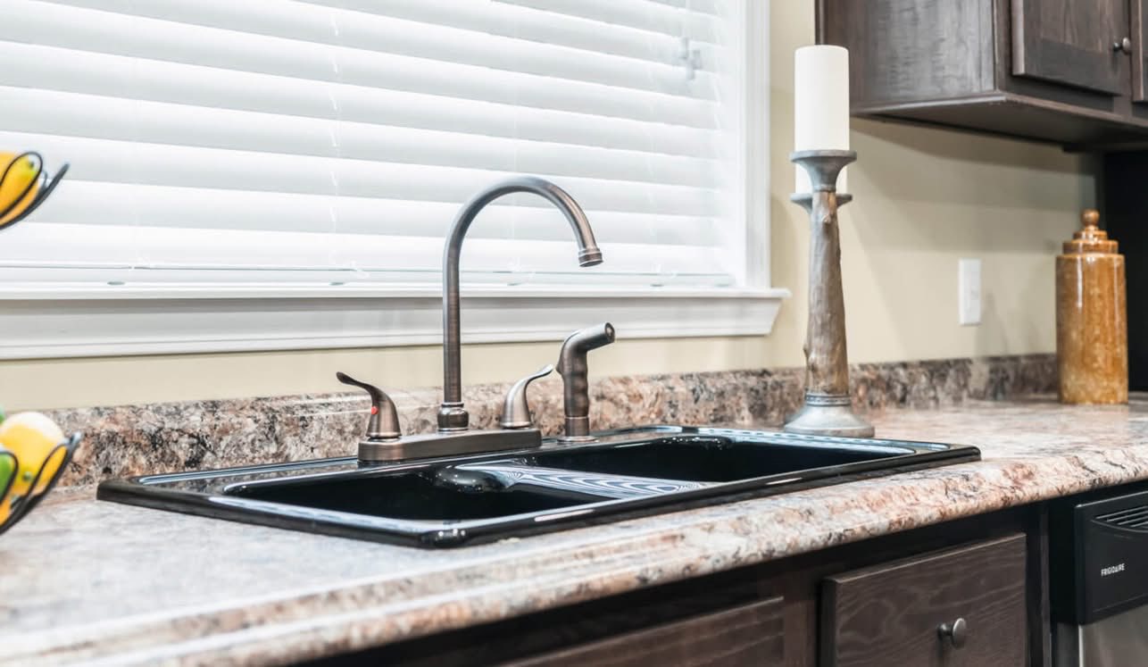 Modern kitchen with a sleek black double sink, silver faucet, and marble countertop. Features a candlestick and decorative jar near a window with blinds.