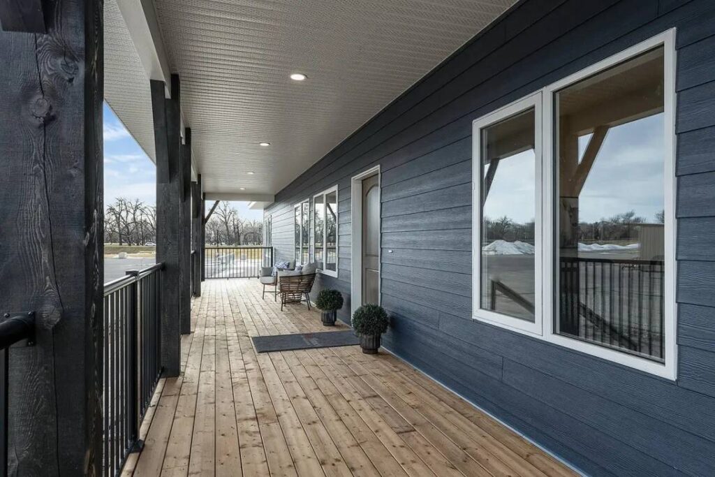 Spacious wooden porch with a modern blue exterior, featuring large windows, potted plants, and seating. The setting is bright and welcoming with a clear sky.