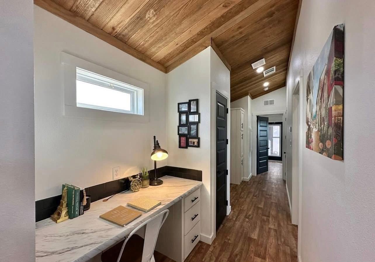 Cozy hallway with wooden ceiling features a marble-topped desk on the left, lit by a lamp. Decor, books, and wall art create a warm, inviting atmosphere.