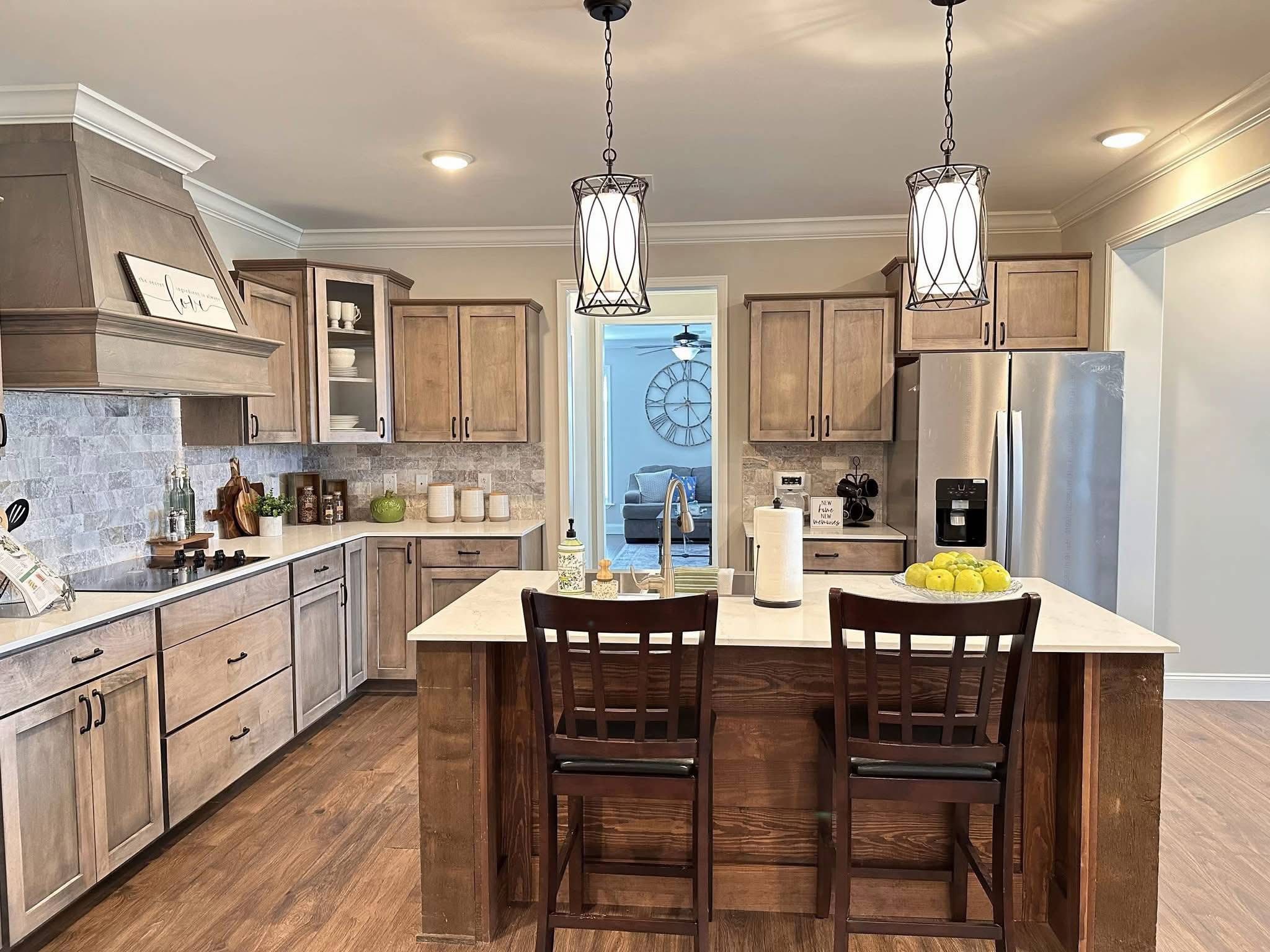 Warm, inviting kitchen with wooden cabinets, marble island, and bar stools. Pendant lights add a cozy ambiance. Stainless steel fridge and decorative items enhance elegance.