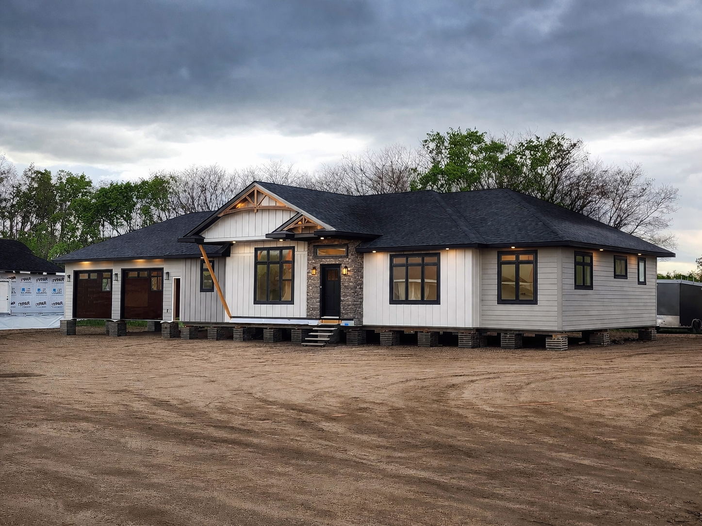 Single-story, modern house under cloudy sky, featuring white siding, black trim, large windows, and a stone entrance. The yard is barren earth.