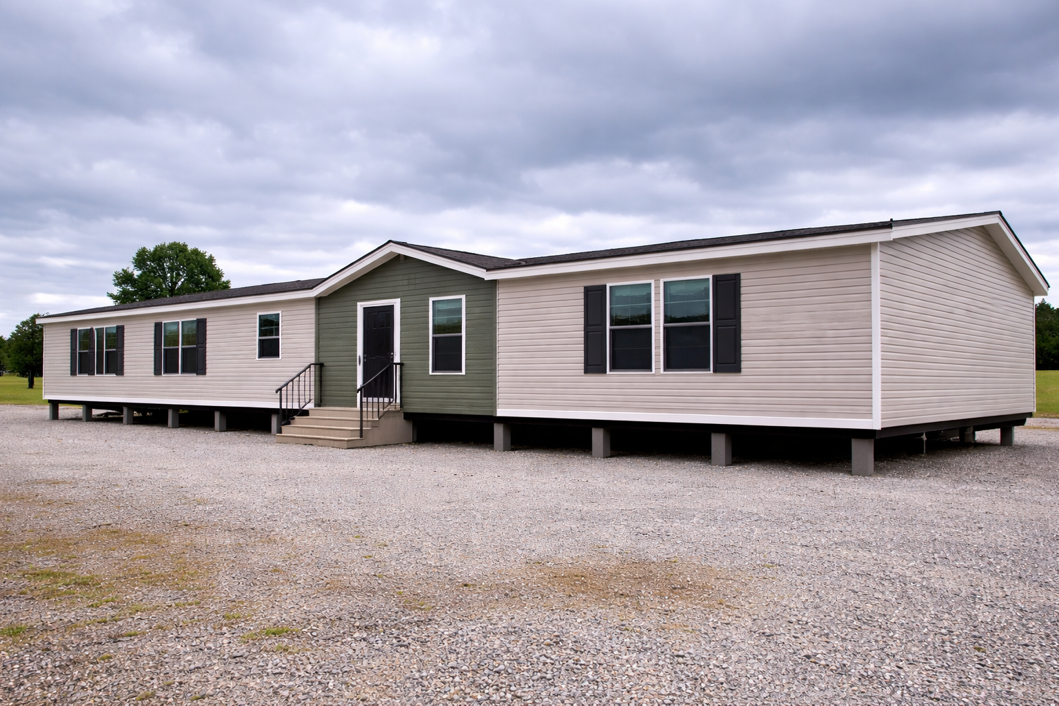 A modern, beige and green manufactured home with black window shutters sits on a gravel lot, under a cloudy sky, conveying a sense of stability and simplicity.