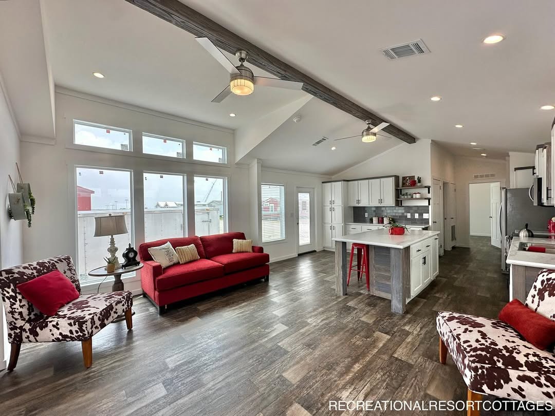 Spacious modern living area with vaulted ceiling, featuring a red couch, cowhide-patterned chairs, large windows, and an open kitchen with white cabinets.