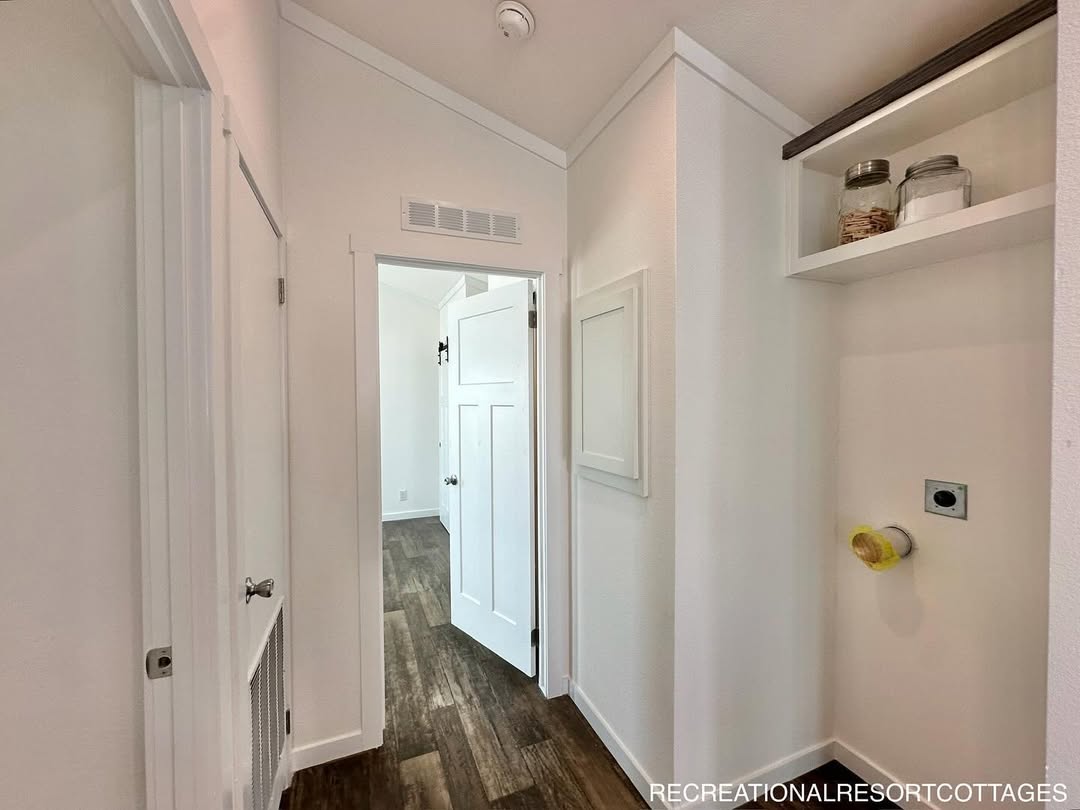 A narrow hallway with white walls and dark wood flooring leads to an open white door. Shelves hold jars on the right, emitting a clean, minimalist vibe.